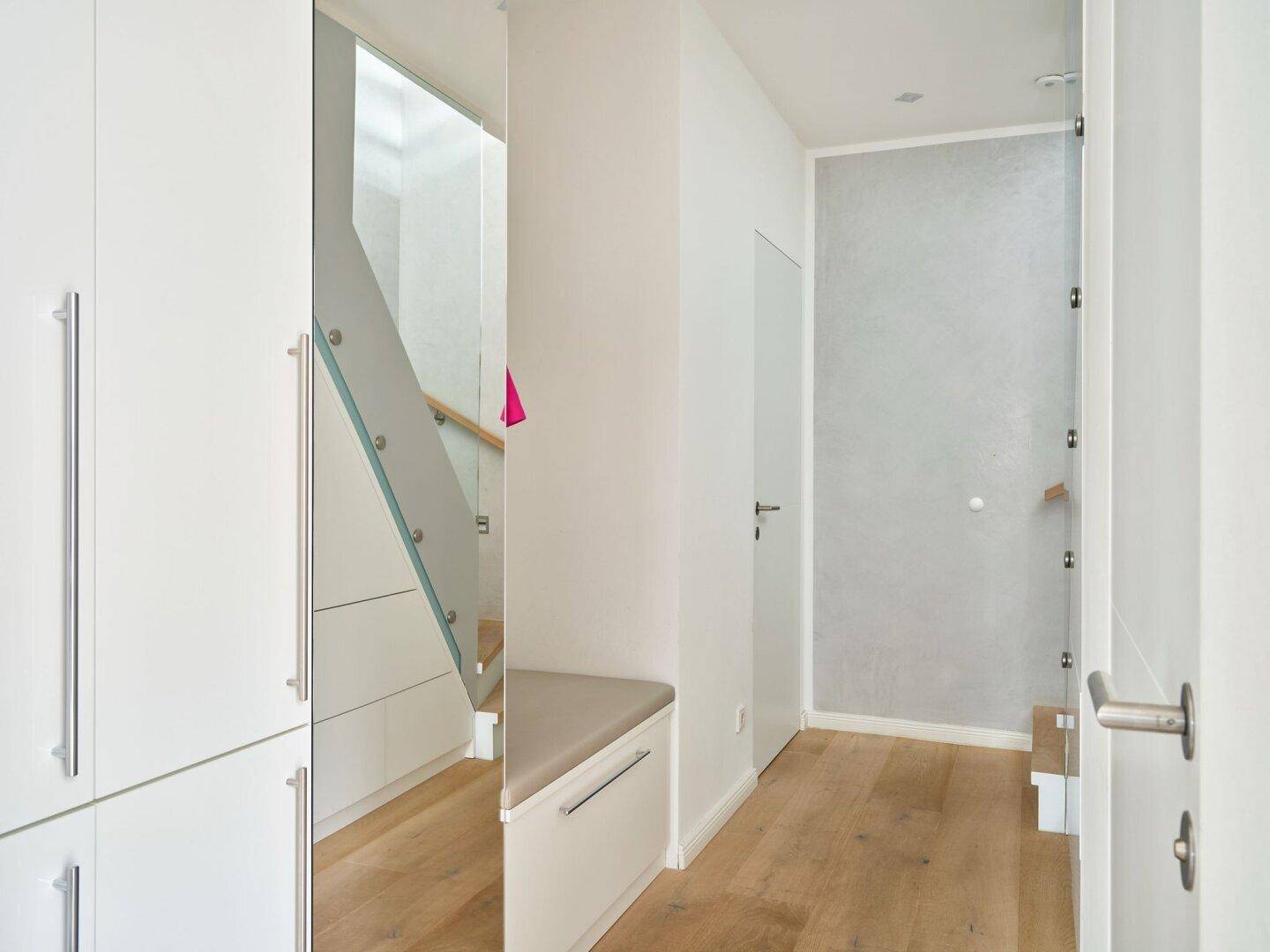 Heller Flur mit Einbauschränken, Sitzbank und Treppe mit Glasgeländer.