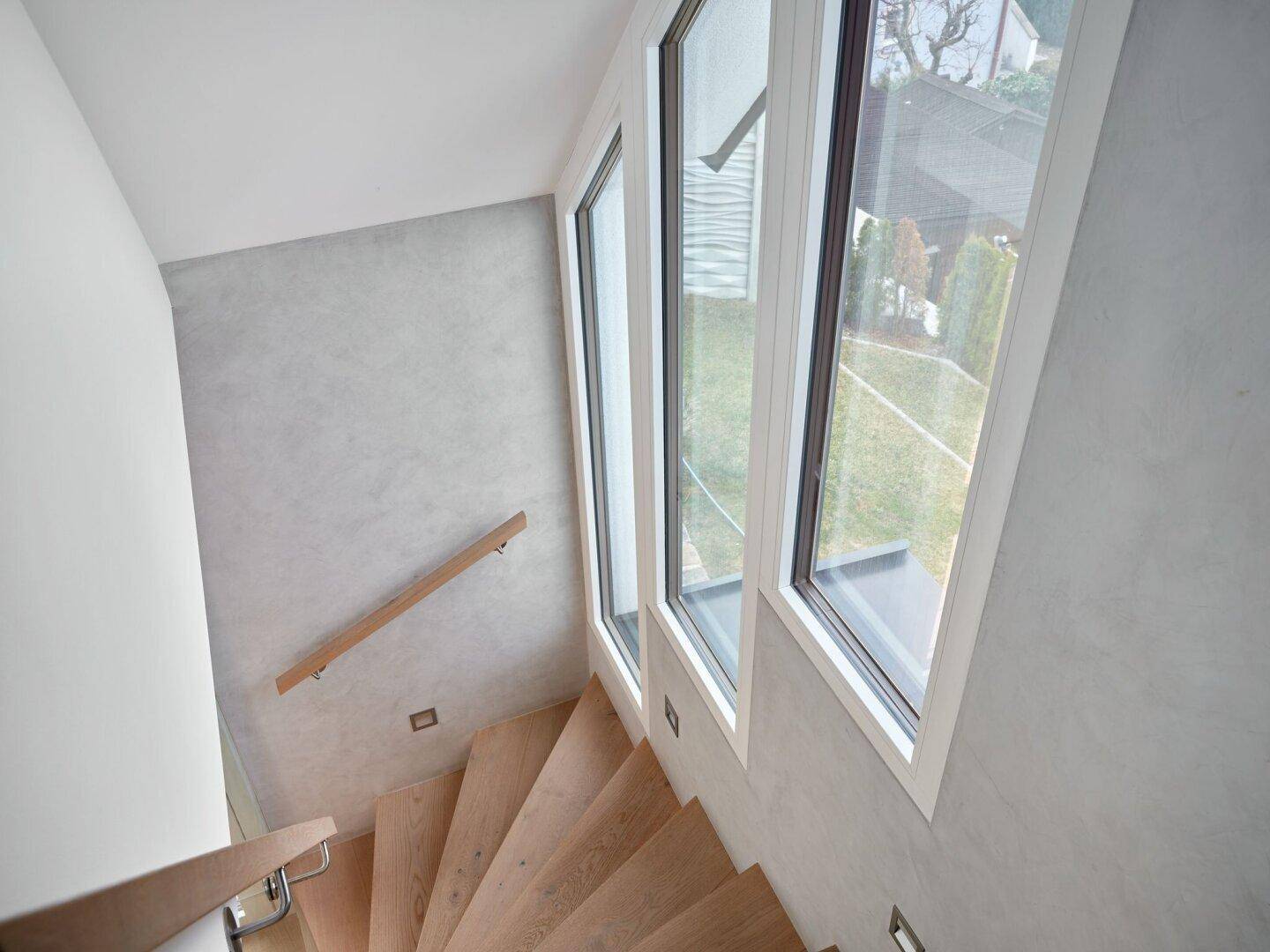 Moderne Holztreppe mit Glasgeländer und großen Fenstern, die viel Licht hereinlassen.