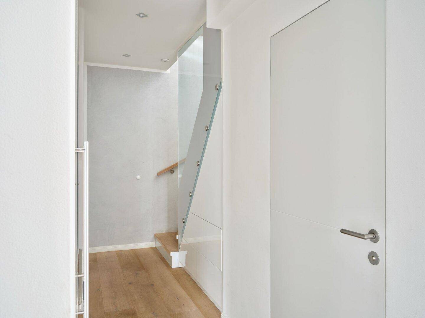 Heller Flur mit moderner weißer Tür und elegantem Glasgeländer, das zu den oberen Etagen führt.