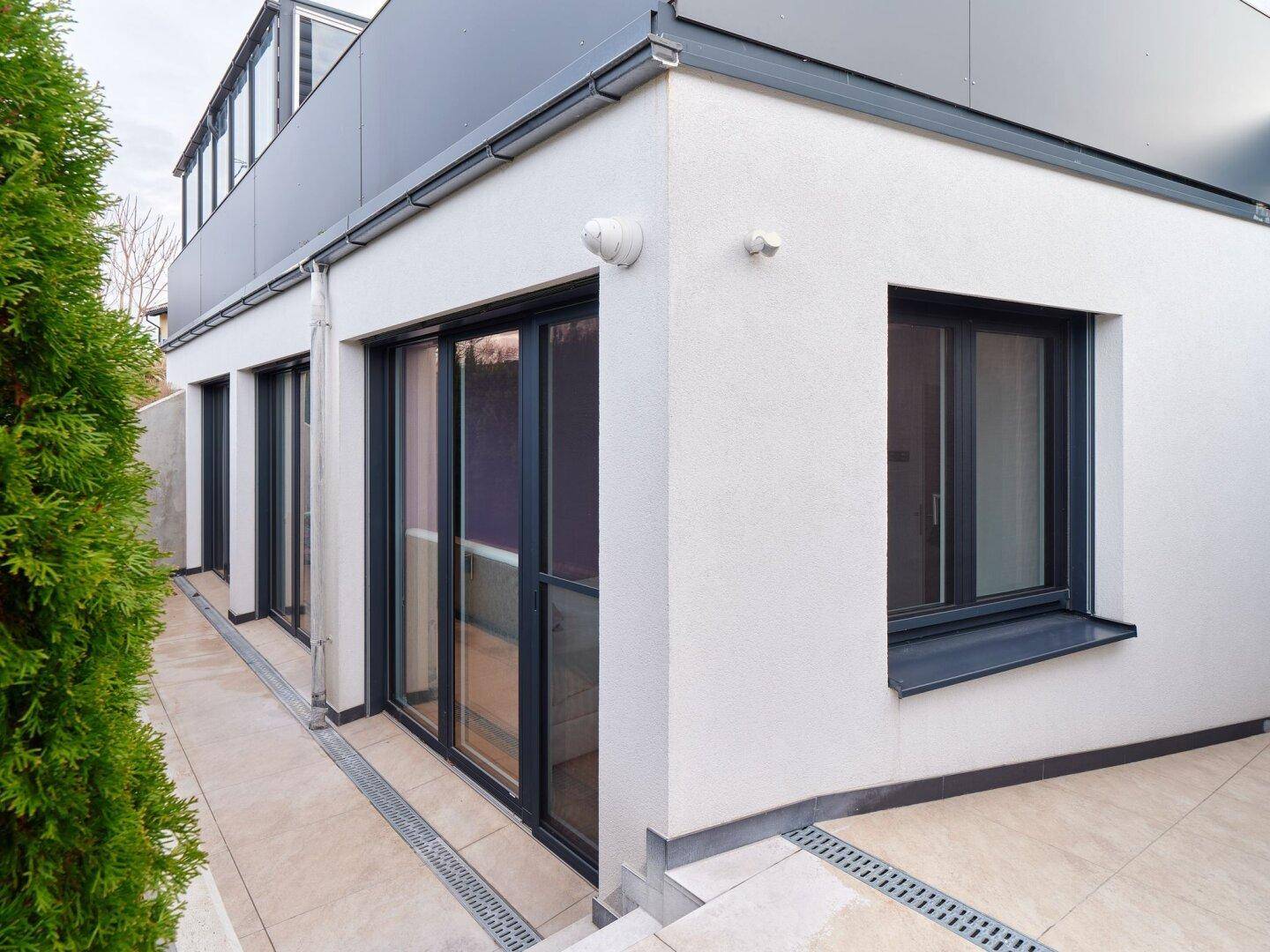 Außenansicht eines modernen Gebäudes mit großer Terrasse und bodentiefen Fenstern, umgeben von Grün.