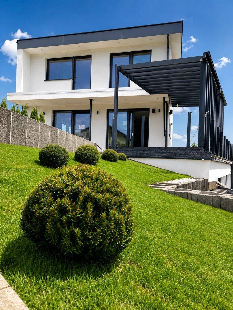 Modernes Haus mit schräger Rasenfläche und dekorativen Büschen unter einer Pergola.