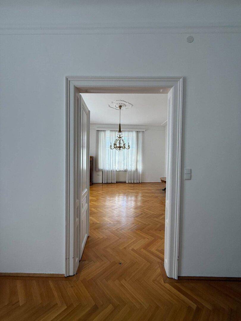 Blick durch eine Tür in ein helles Zimmer mit Fischgrätparkett und Stuckdecke.