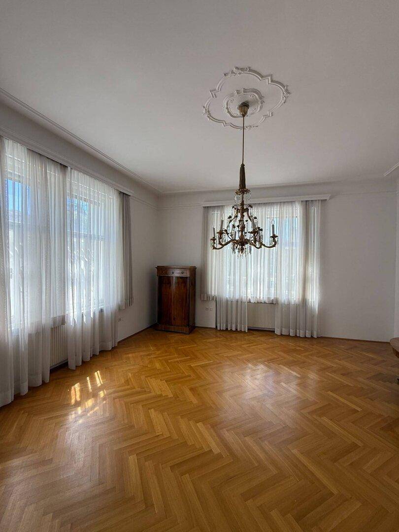 Helles Wohnzimmer mit Fischgrätparkett, großen Fenstern und klassischem Kronleuchter.