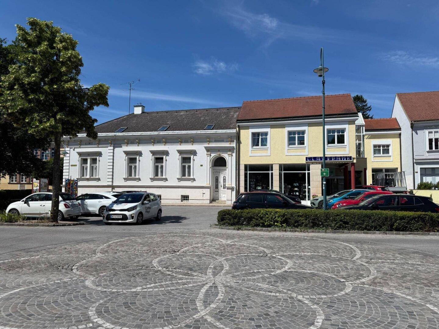 Straßenansicht mit historischen Gebäuden, geparkten Autos und einem gepflasterten Platz.