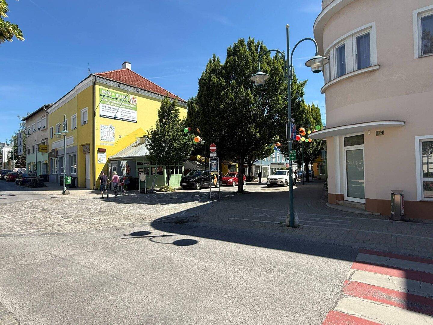 Belebtes Stadtbild mit Geschäften, Bäumen und einer Straße unter blauem Himmel.
