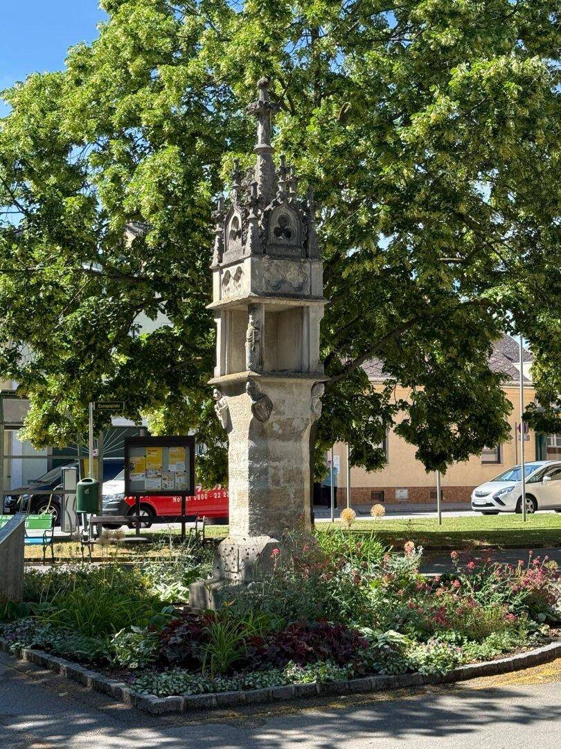 Historisches Steindenkmal in einer kleinen Grünanlage mit Bäumen im Hintergrund.