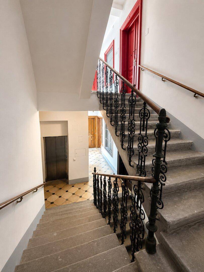 Historisches Treppenhaus mit schmiedeeisernem Geländer, roten Türen und Aufzug im Hintergrund.