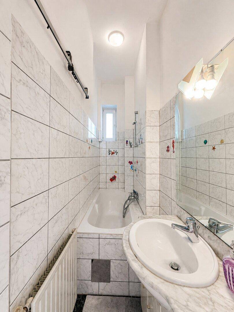Schmaler Badezimmerbereich mit Badewanne, Waschbecken, Spiegel und weißen Fliesenwänden.
