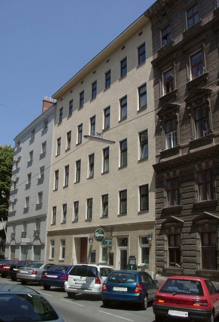 Mehrstöckiges Wohngebäude an einer Straße mit geparkten Autos und Geschäften im Erdgeschoss.