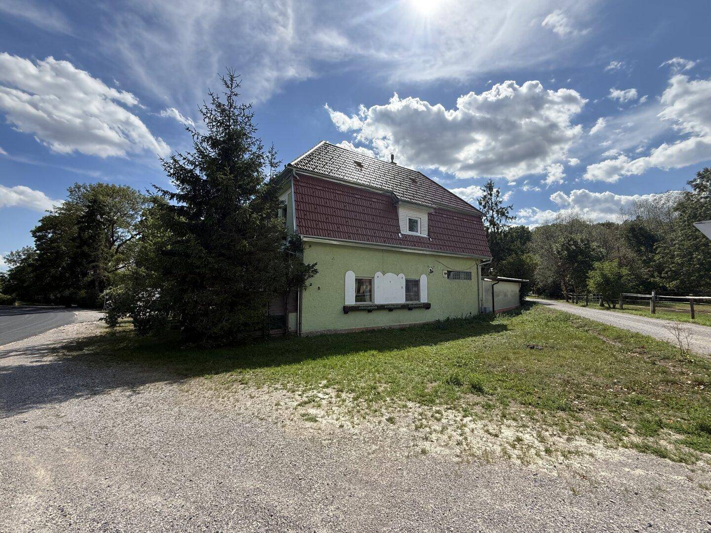 Seitenansicht eines älteren Hauses mit grüner Fassade, roten Dachziegeln und umgebender Natur.