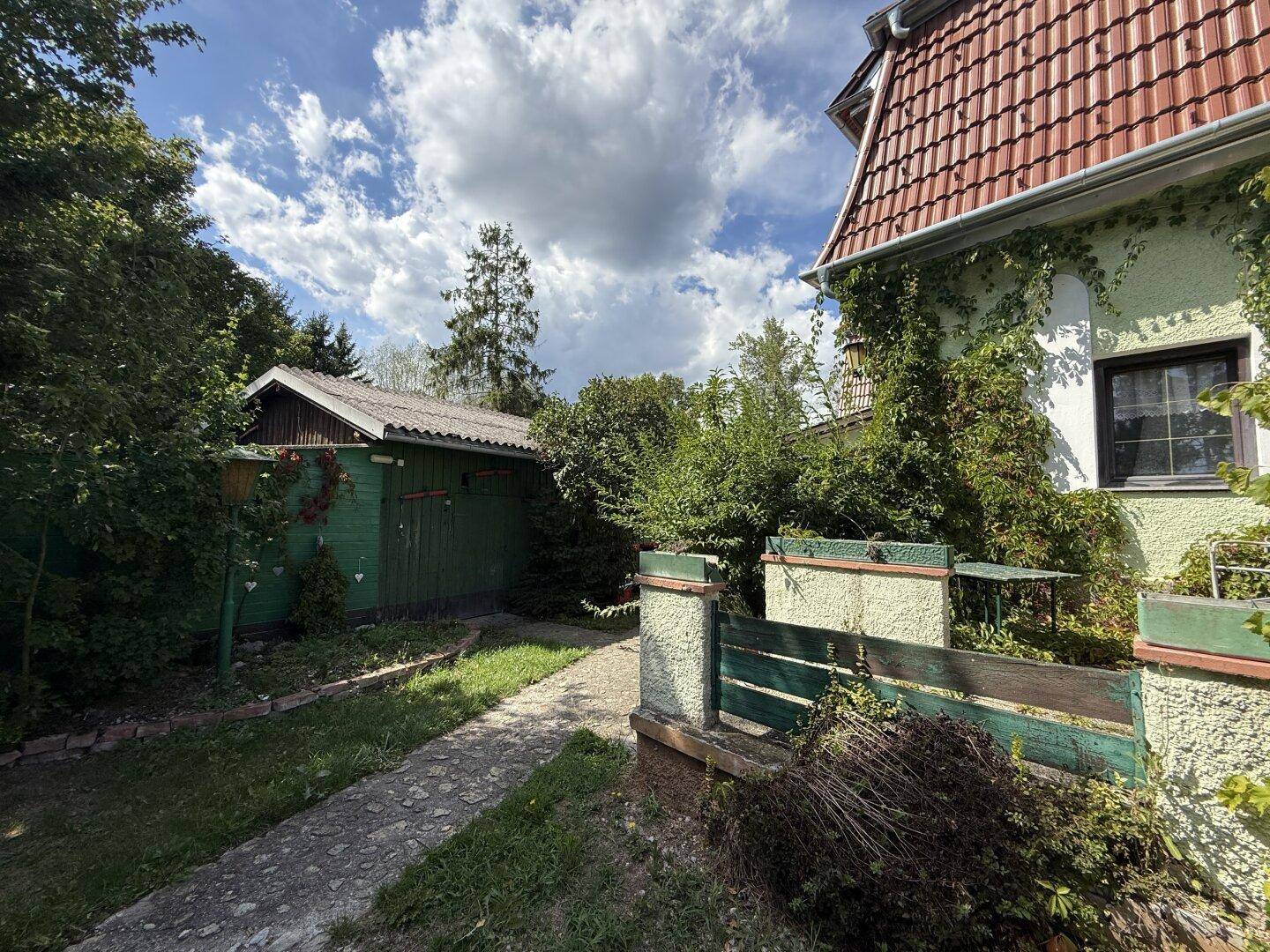 Grüner Schuppen im Gartenbereich neben dem Haus, umgeben von Bäumen und einem Pfad.