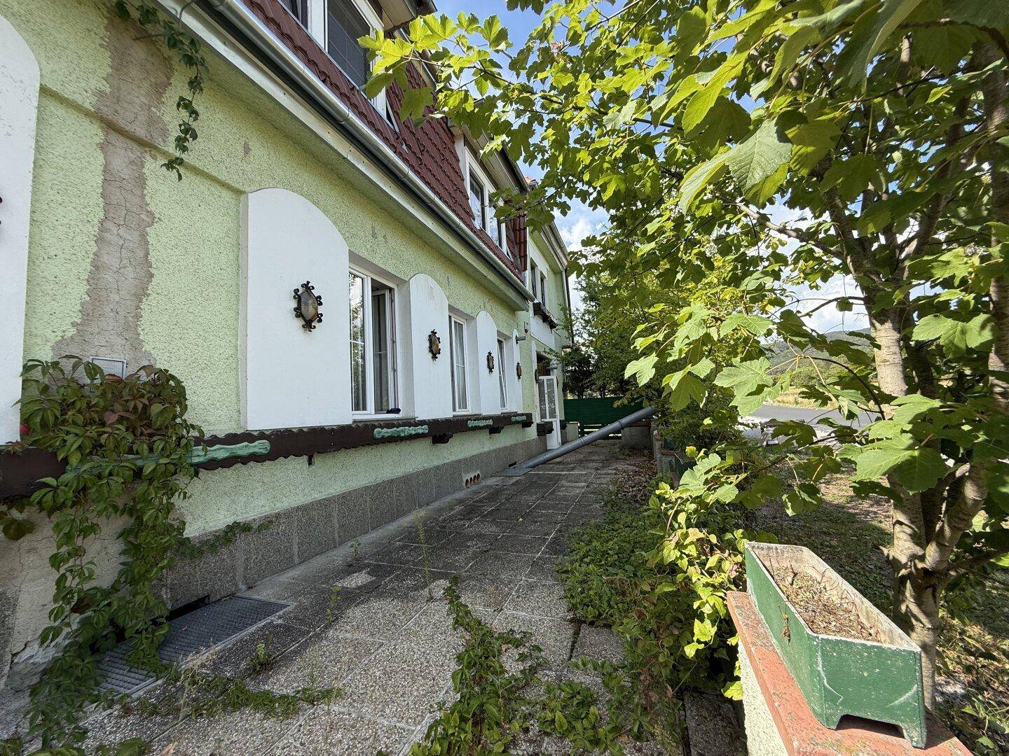 Langer Weg entlang der grünen Hausfassade mit Fenstern und Bäumen im Hintergrund.
