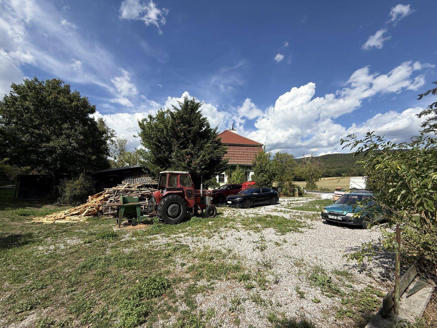 Großes Grundstück mit einem Traktor, Holzstapeln und geparkten Autos vor dem Haus.