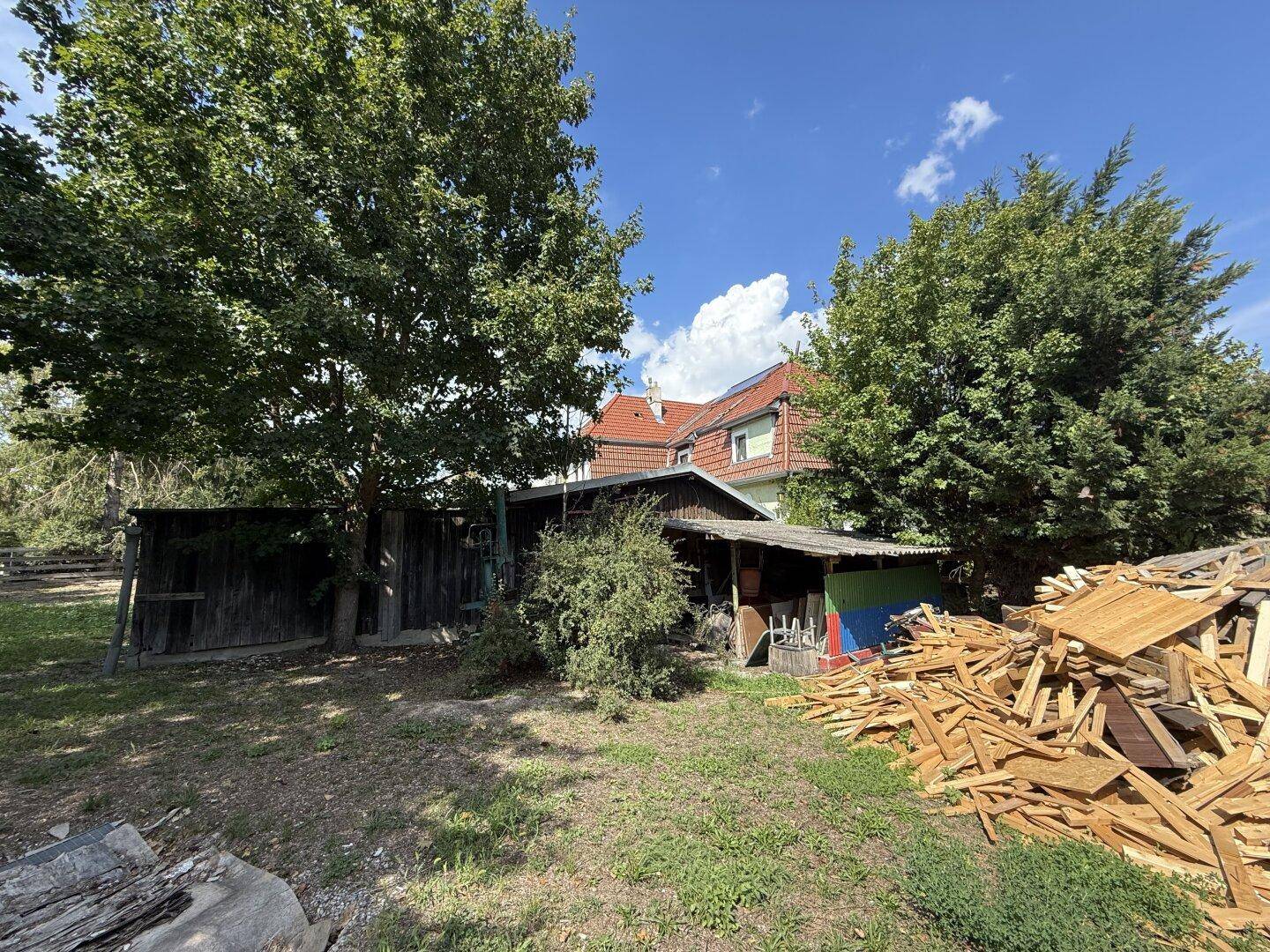 Alter Schuppen und große Holzstapel im Gartenbereich, mit dem Haupthaus im Hintergrund.