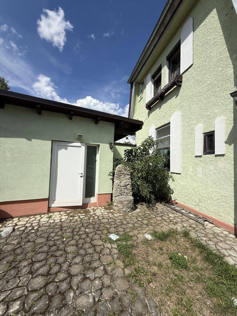 Gepflasterter Bereich neben einem Anbau mit weißer Tür und Steinmauer.
