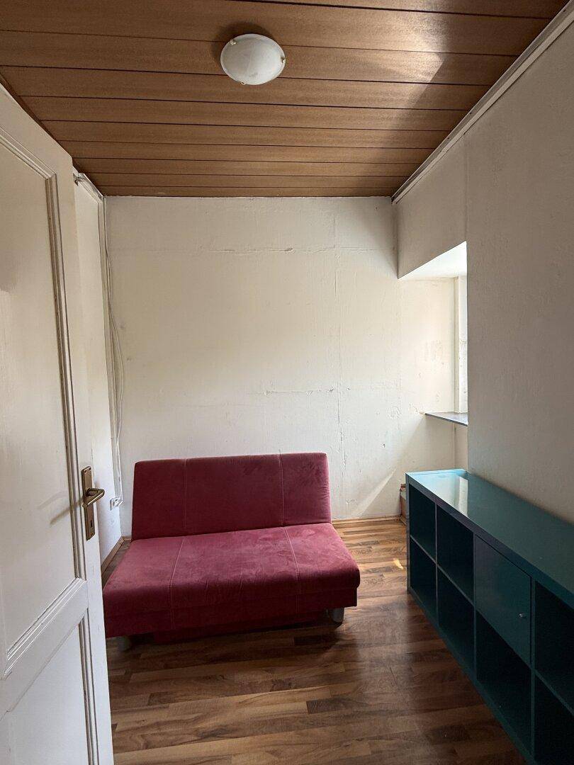 Kleines Zimmer mit rotem Schlafsofa, Holzdecke und Fenster, nutzbar als Schlaf- oder Gästezimmer.