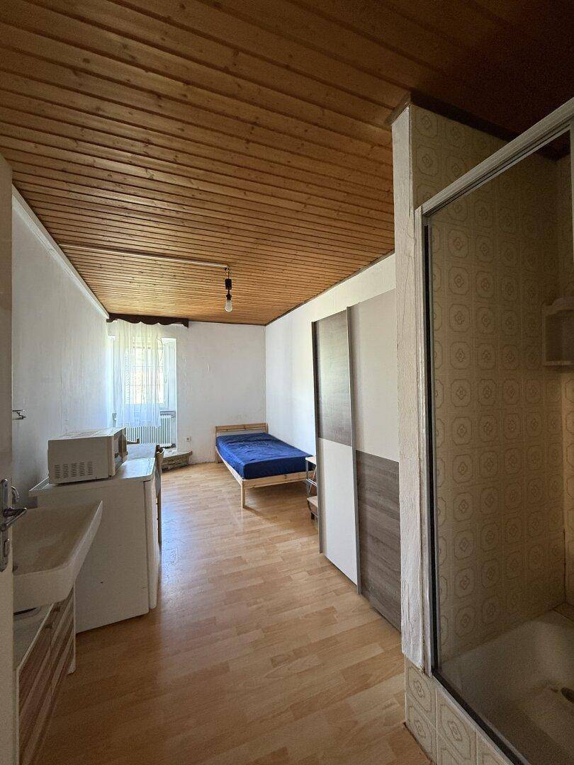 Schlafzimmer mit Bett, Waschbecken, Mikrowelle und Duschkabine, mit Holzdecke und Laminatboden.