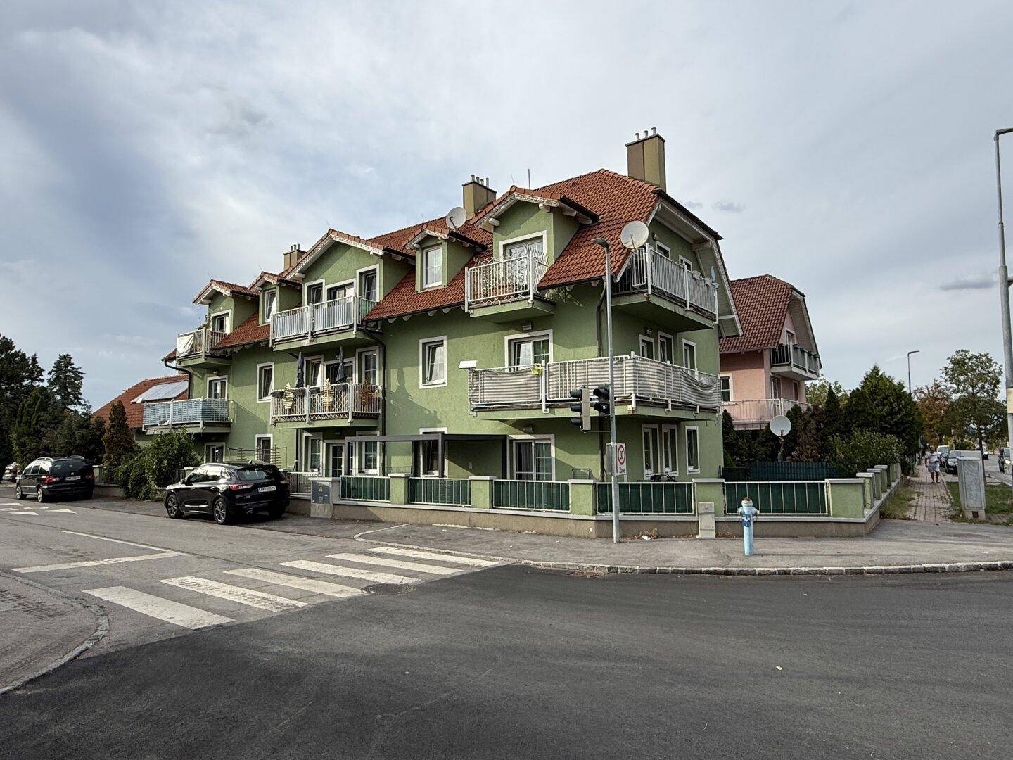 Mehrfamilienhaus mit grüner Fassade, roten Ziegeldächern und mehreren Balkonen an einer Straßenecke.
