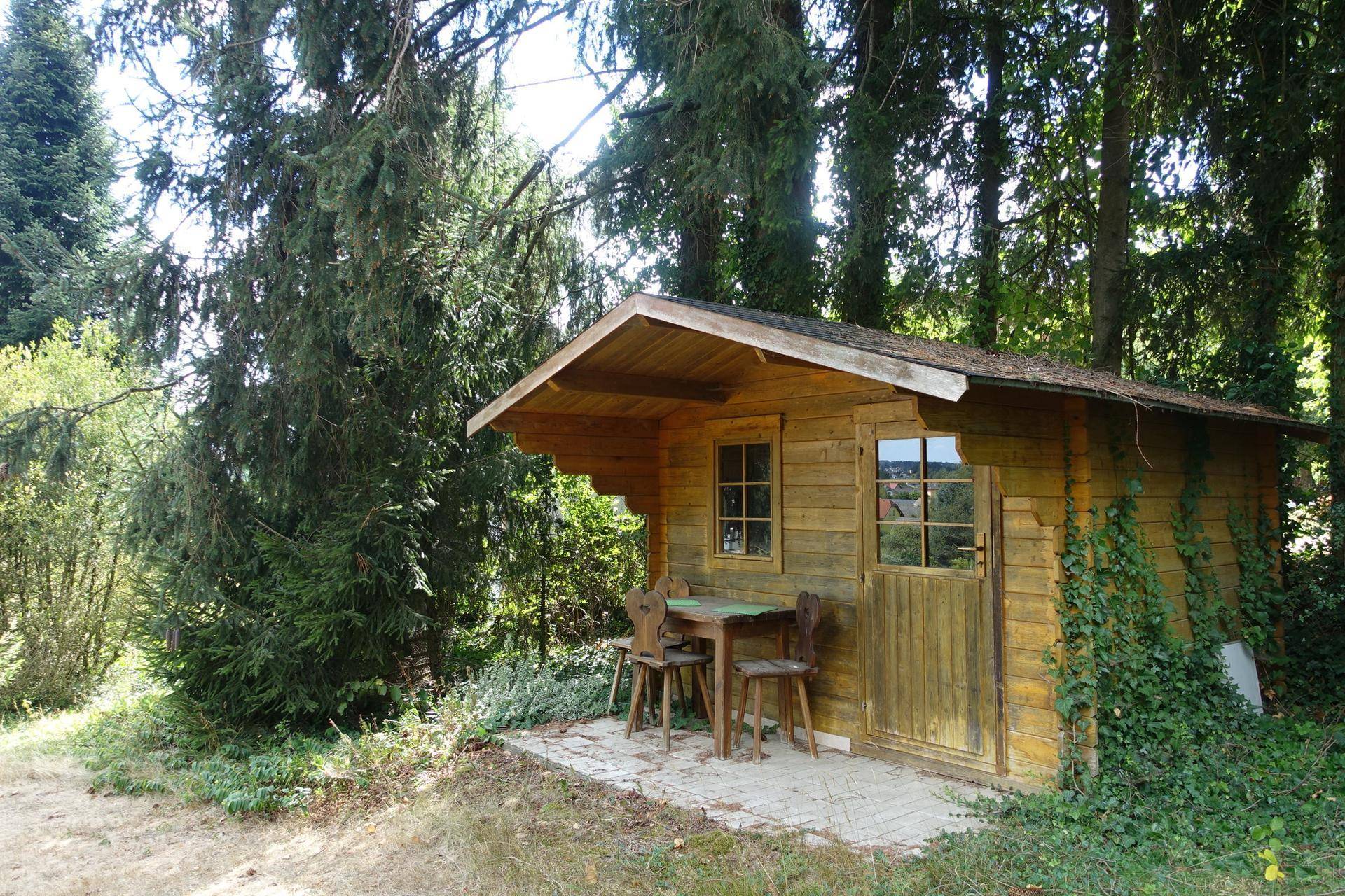 Gemütliches Gartenhaus aus Holz mit kleiner Terrasse und Sitzgelegenheiten, umgeben von Bäumen.