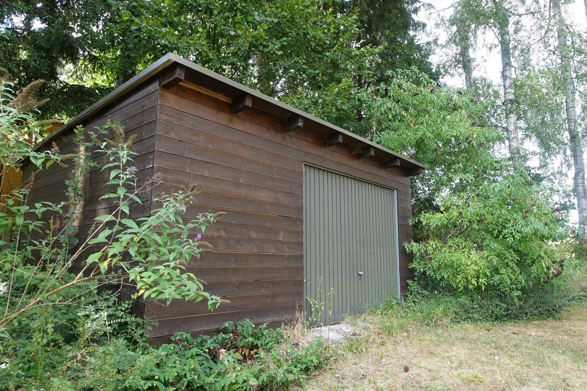 Funktionales Holzgartenhaus mit einem grünen Tor, eingebettet in eine grüne Umgebung mit Bäumen.