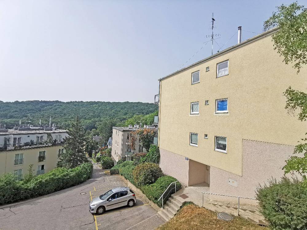 Außenansicht eines Wohngebäudes mit Parkplatz und Blick auf bewaldete Hügel.