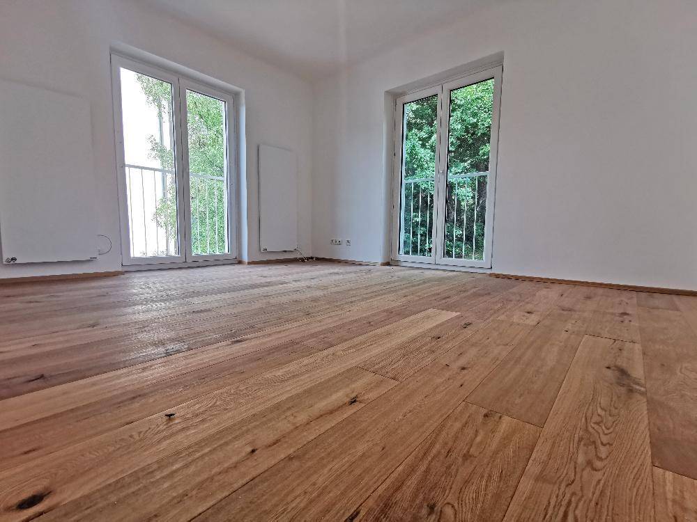 Geräumiges Zimmer mit hellem Holzboden und Zugang zu zwei französischen Balkonen mit grüner Aussicht.