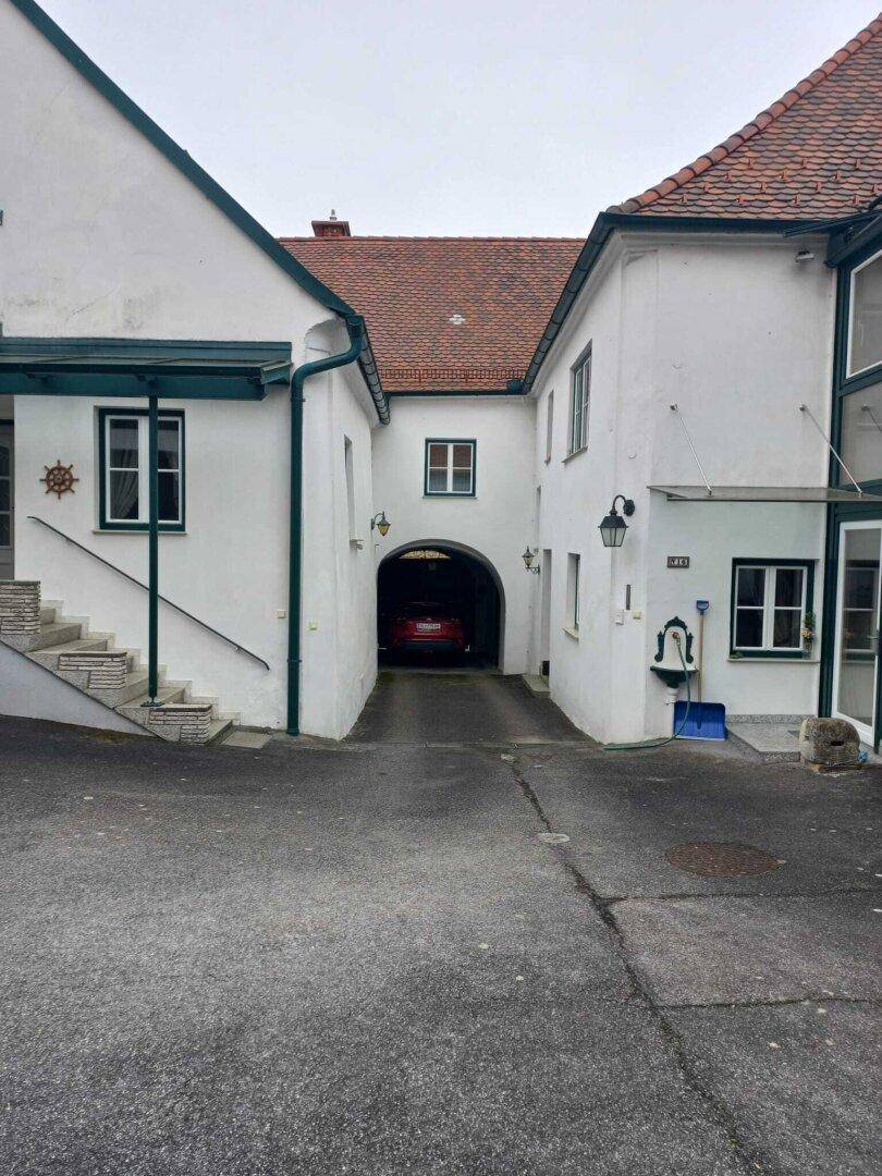Einfahrt zu einem Innenhof mit einem Torbogen, der zu einem überdachten Parkplatz führt.