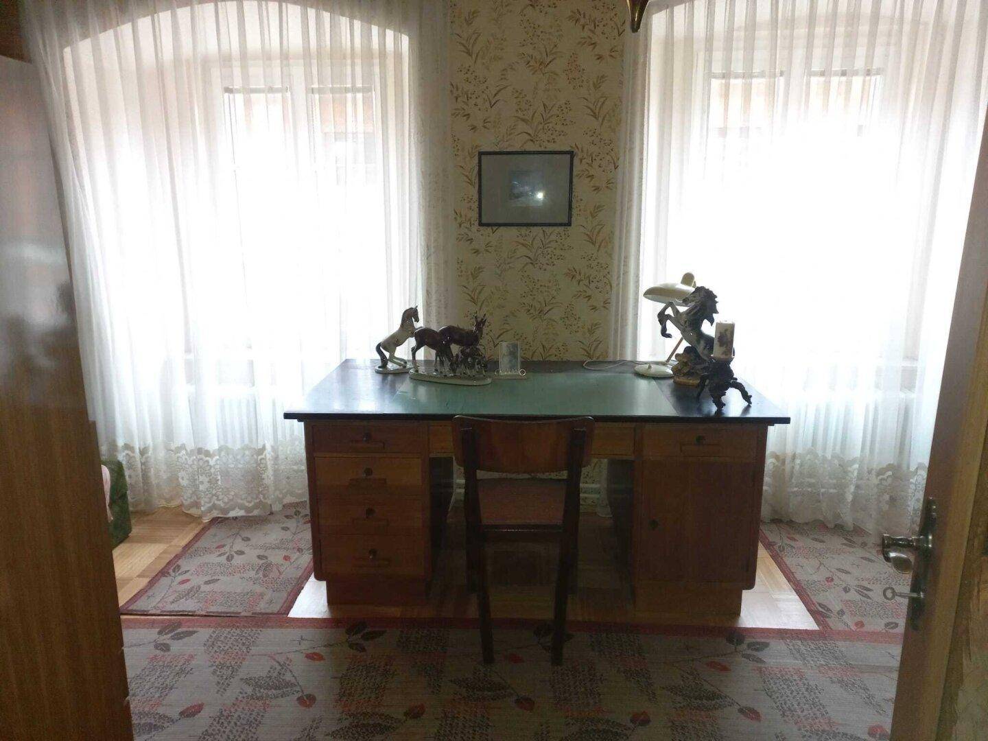 Helles Arbeitszimmer mit großem Holzschreibtisch, gemusterter Tapete und Blick nach draußen.