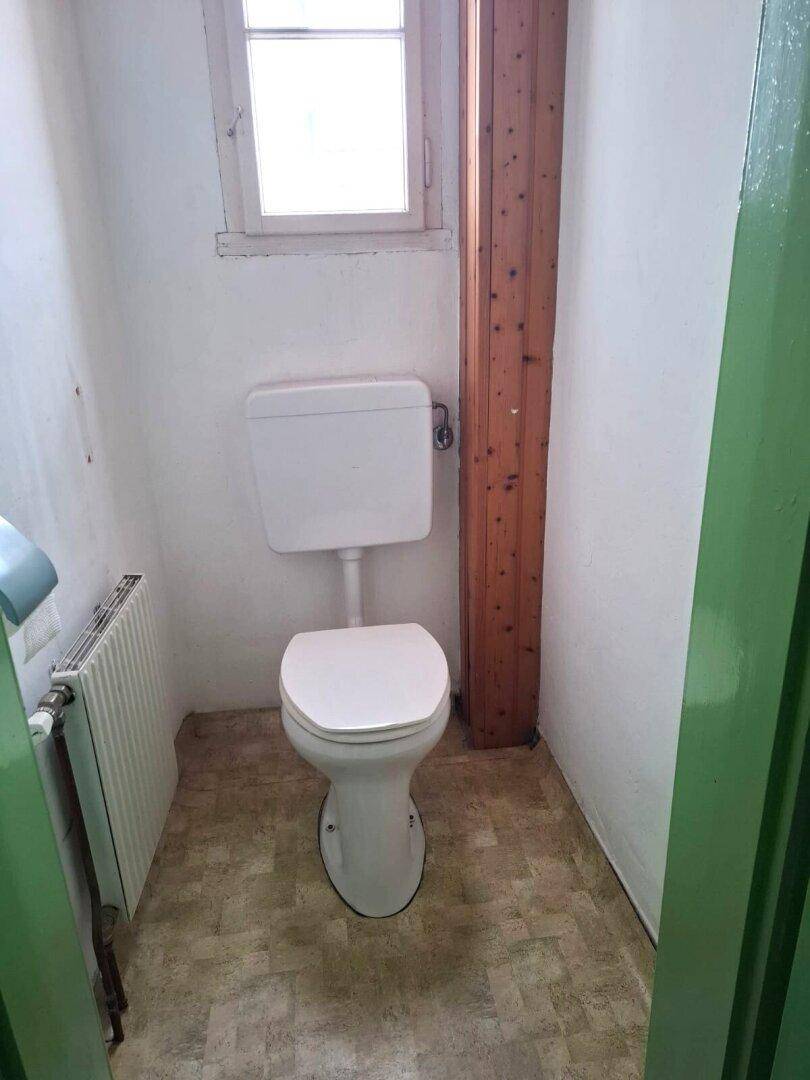 Einfaches WC mit Fenster, Heizkörper und grüner Tür in einem kleinen, funktionalen Raum.
