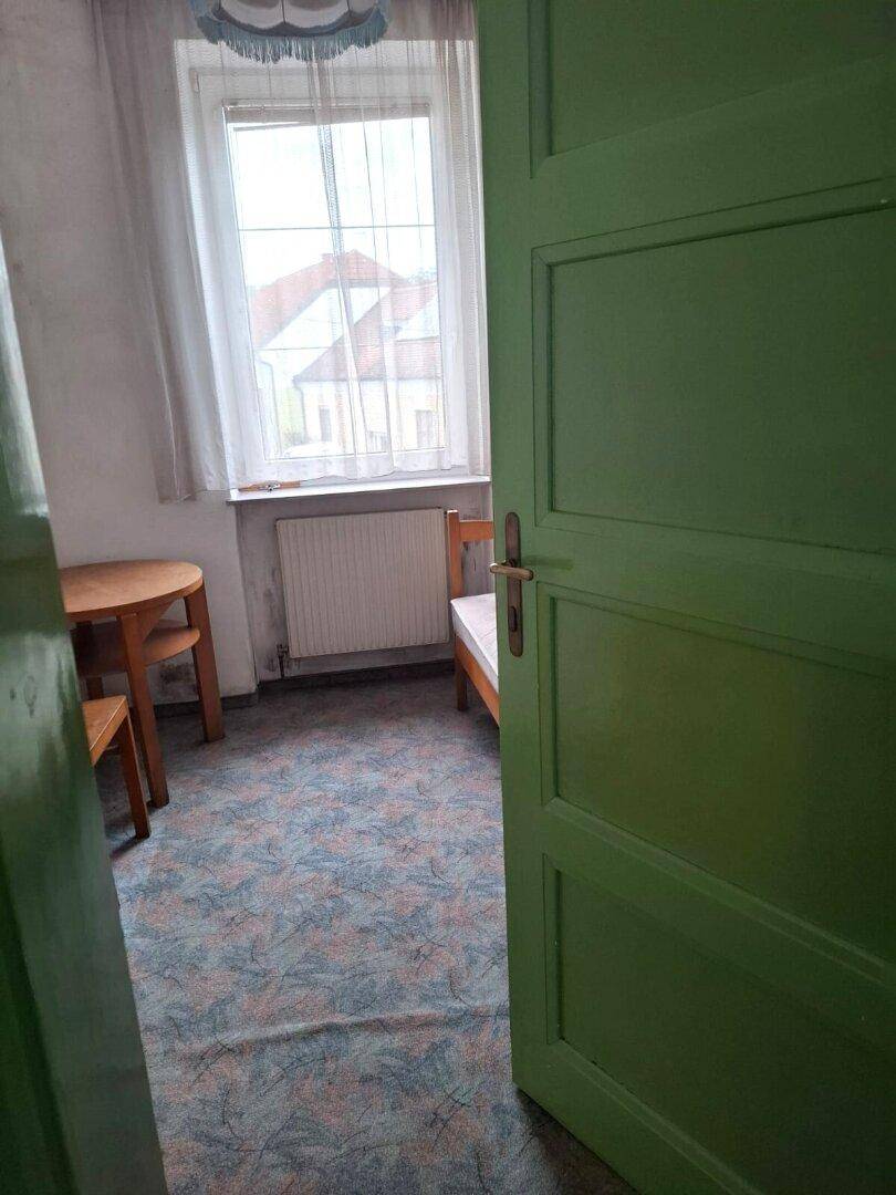 Kleines Zimmer mit gemustertem Teppichboden, Fenster und einem einfachen Tisch mit Stühlen.