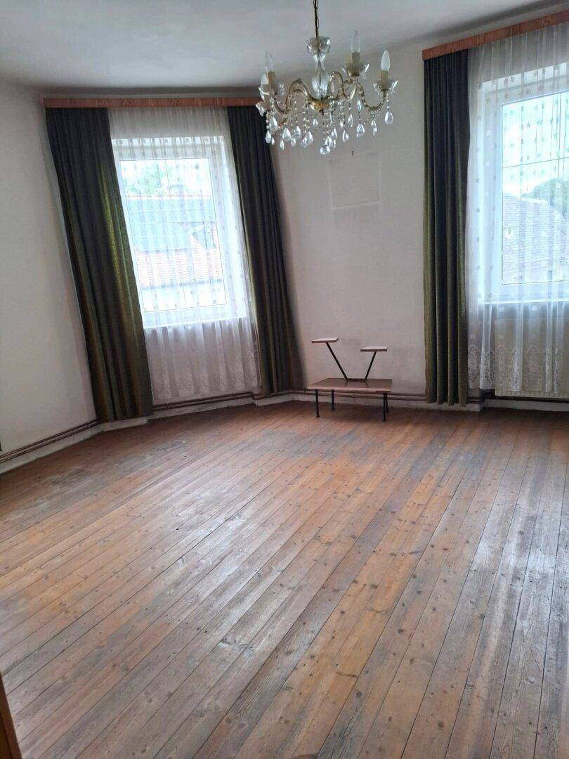 Geräumiges Wohnzimmer mit Holzdielenboden, zwei großen Fenstern und einem eleganten Kronleuchter.