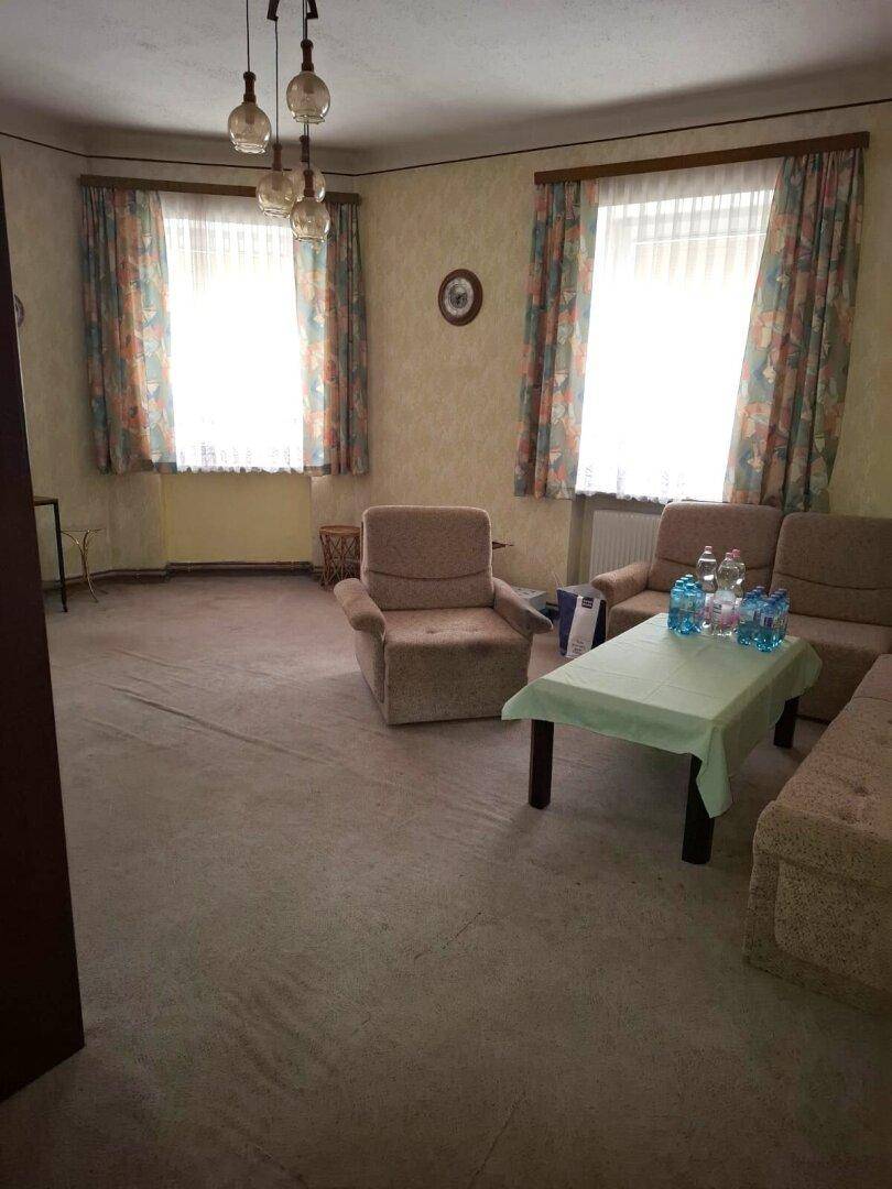 Wohnzimmer mit Teppichboden, zwei Sesseln, einem Sofa und einem Couchtisch mit Wasserflaschen.