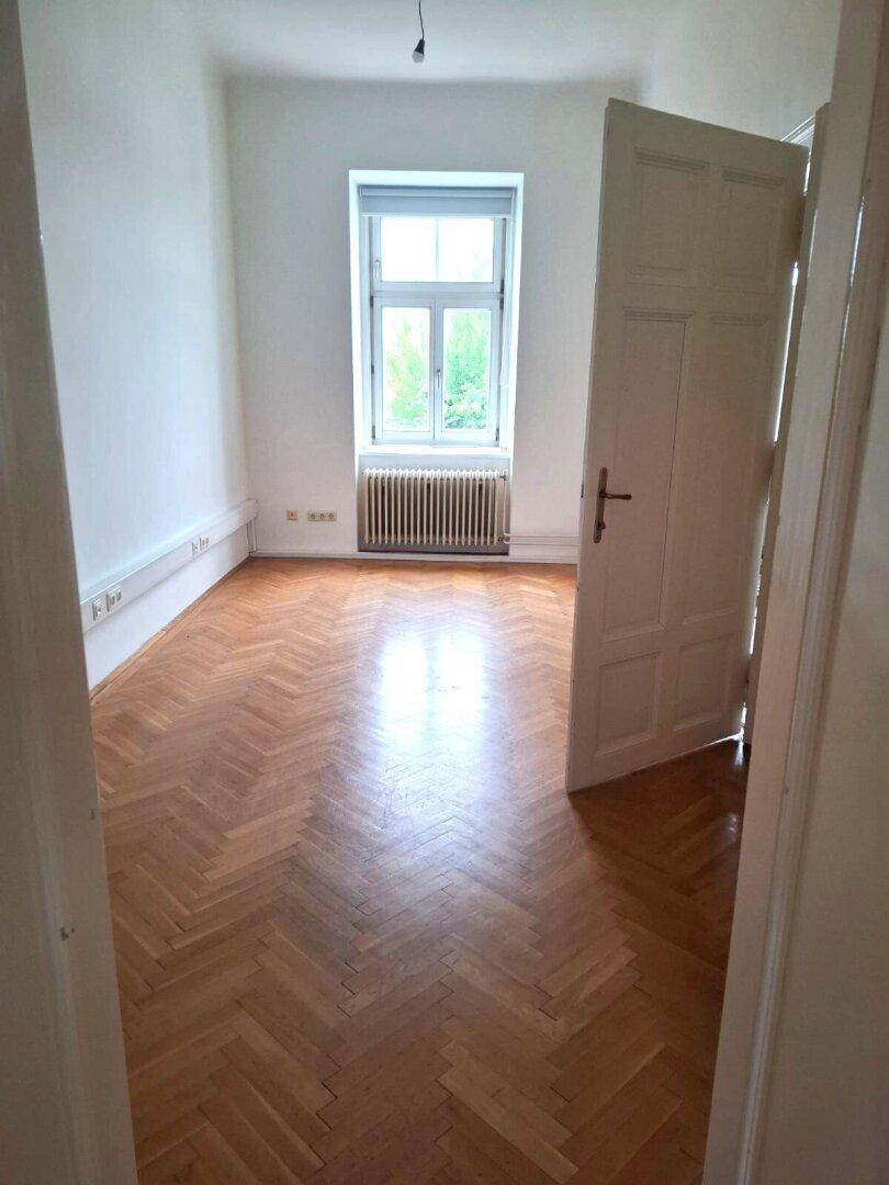 Leeres Zimmer mit glänzendem Fischgrätparkettboden, einem Fenster und einem Heizkörper, bereit zur Einrichtung.