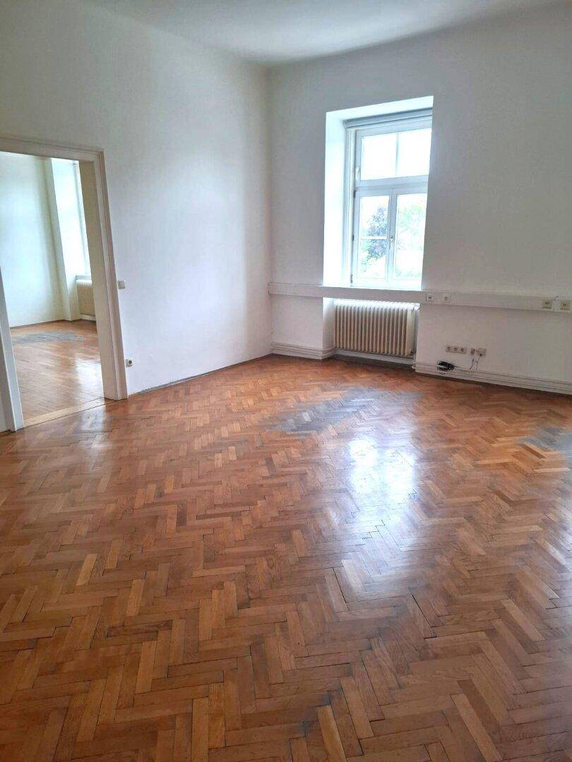 Leerer Raum mit glänzendem Fischgrätparkettboden, einem Fenster und einem Heizkörper, der auf Renovierung wartet.