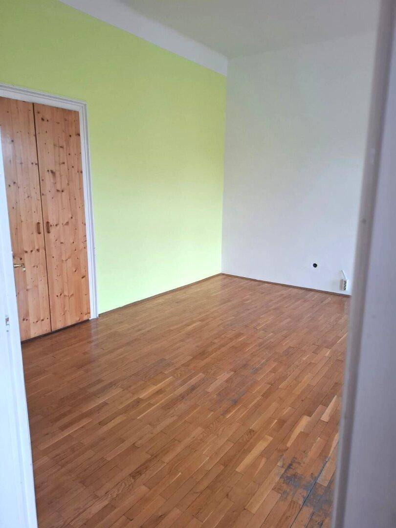 Leeres Zimmer mit Holzboden, einer hellgrünen Akzentwand und einer einfachen Holztür.