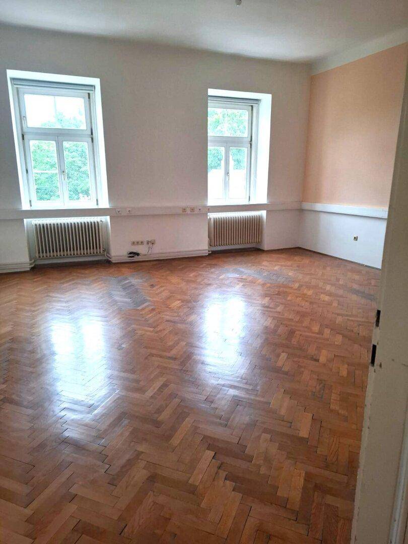 Geräumiges Zimmer mit glänzendem Fischgrätparkettboden, zwei Fenstern und Heizkörpern, bereit zur Gestaltung.