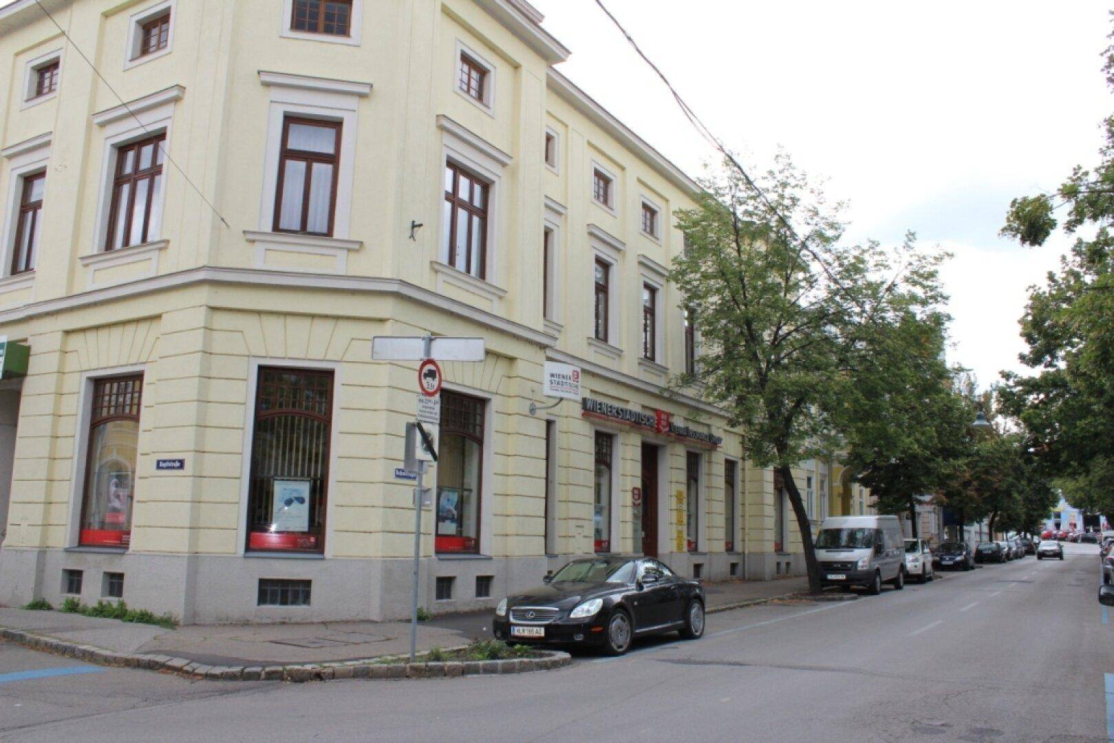 Eckgebäude mit gelber Fassade, großen Fenstern und Blick auf die Straße mit parkenden Autos.