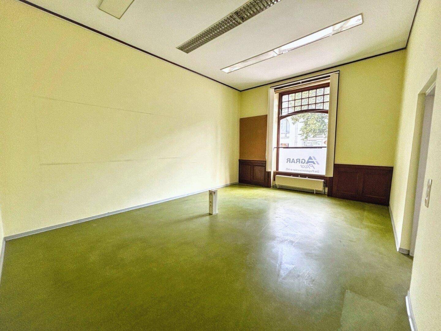 Heller Büroraum mit grünem Boden, gelben Wänden und einem großen Fenster.