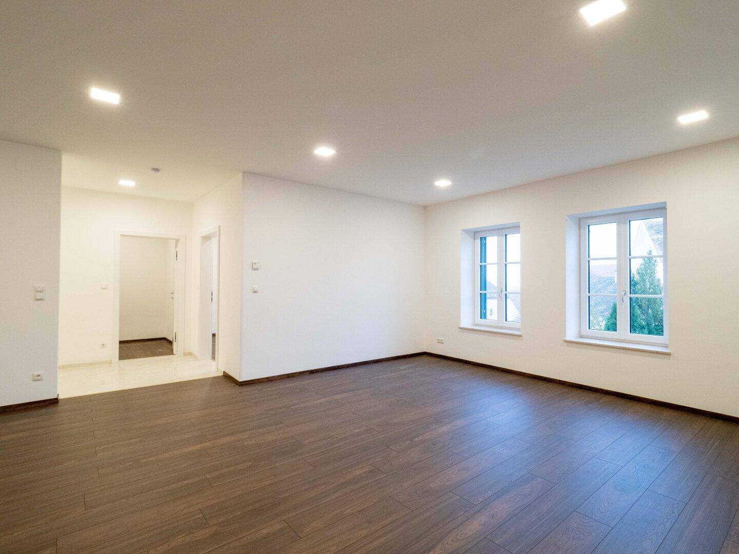 Helles Wohnzimmer mit dunklem Holzboden und zwei Fenstern, das viel Tageslicht hereinlässt.