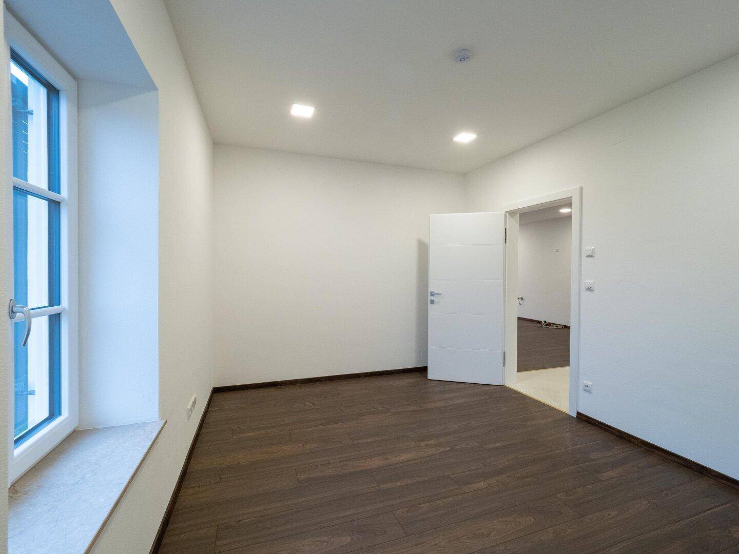 Helles Zimmer mit dunklem Holzboden und einem großen Fenster, ideal als Schlaf- oder Arbeitszimmer.