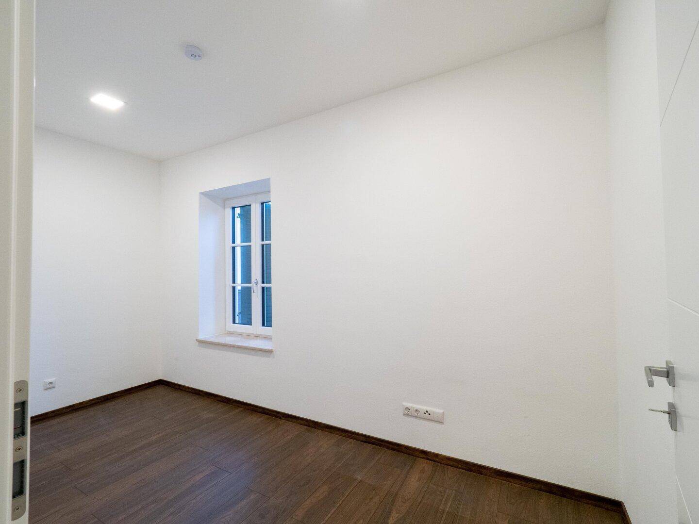 Ruhiges Zimmer mit dunklem Holzboden und einem Fenster, das viel Tageslicht hereinlässt.