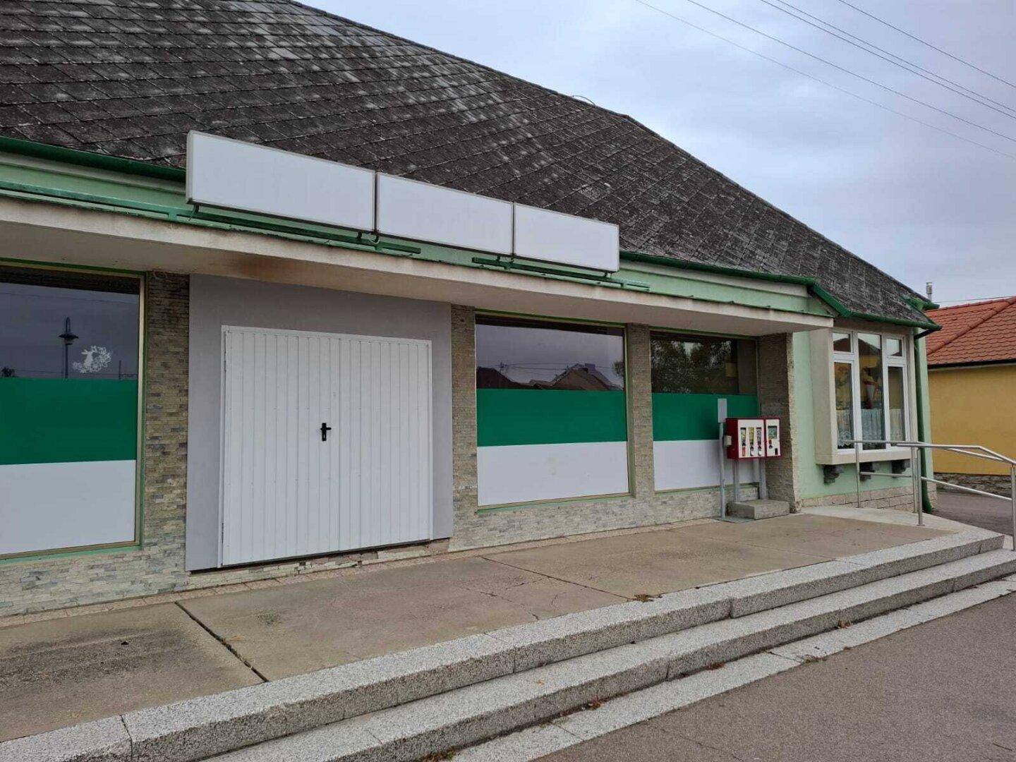 Nahaufnahme der Ladenfront mit Eingangstür, Schaufenstern und Werbeflächen, inklusive einer kleinen Rampe.