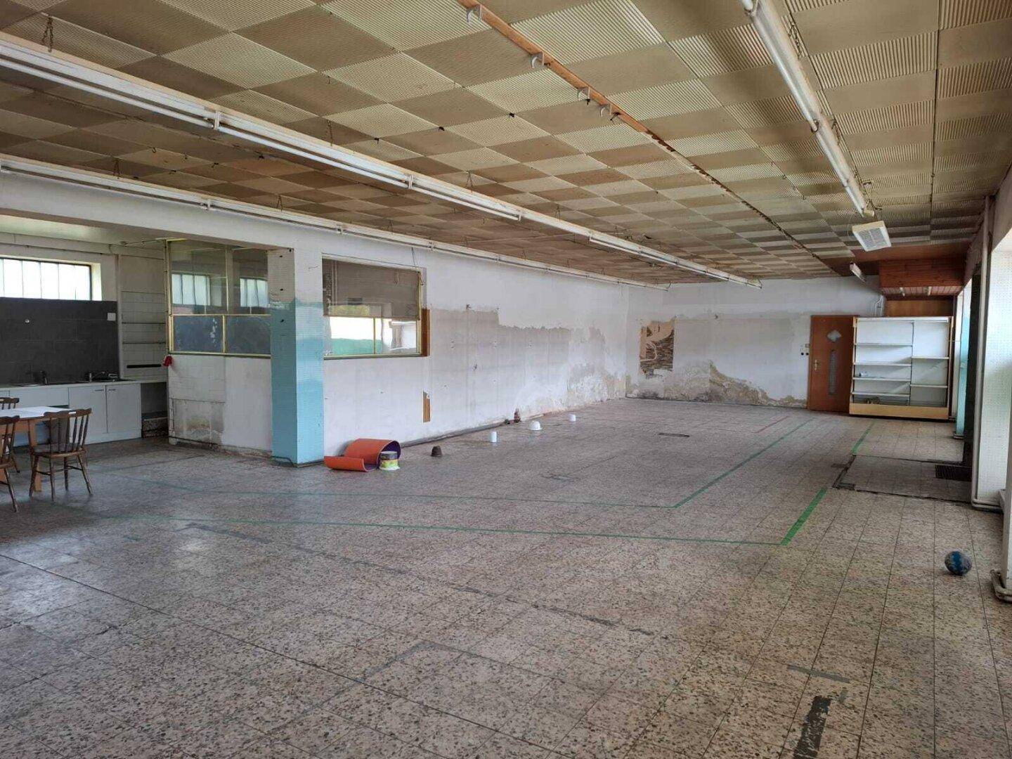 Großer, leerstehender Raum mit Fliesenboden und abgehängter Decke, der deutliche Renovierungsbedürftigkeit aufweist.