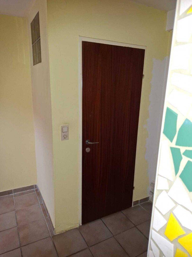 Schmaler Flur mit Fliesenboden und einer Holztür, der einen Übergangsbereich in einem älteren Gebäude darstellt.
