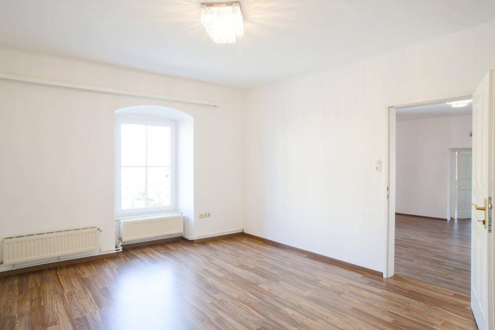 Helles Zimmer mit Holzboden und einem einzelnen Fenster mit Rundbogen.