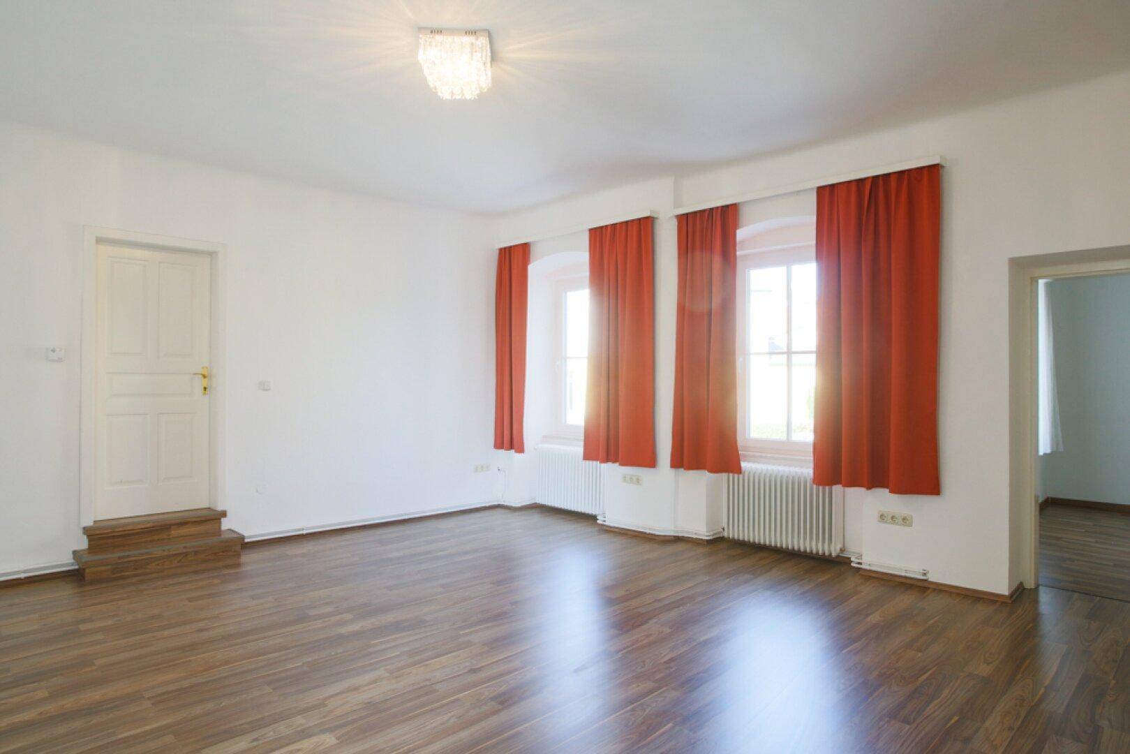 Helles Zimmer mit Holzfußboden, roten Vorhängen und einer Tür mit kleiner Stufe.