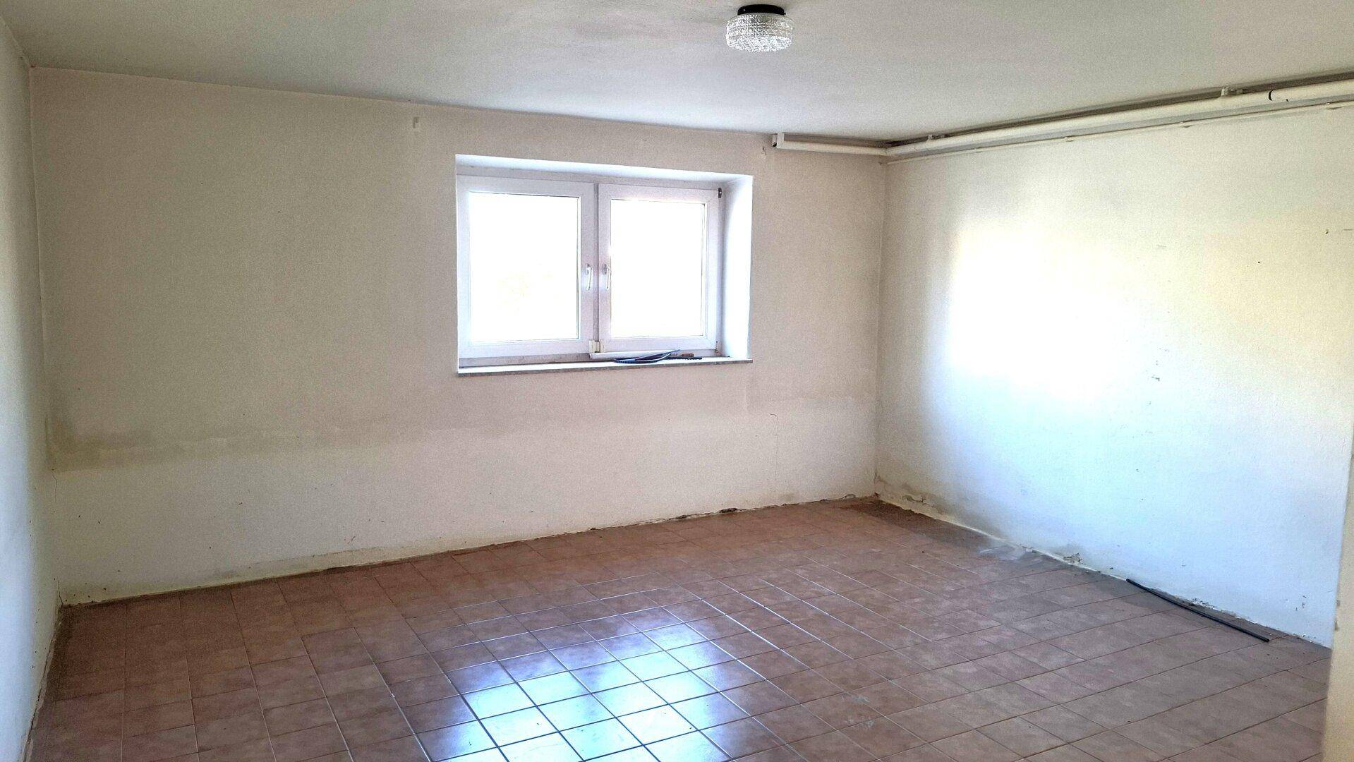 Leerer Raum im Untergeschoss mit Fliesenboden und einem Fenster für Tageslicht.