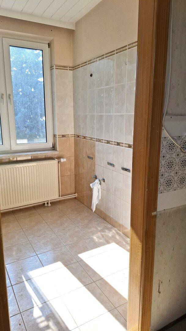 Badezimmer mit Fliesen an Wänden und Boden, Fenster und Heizkörper, bereit für Sanierung.