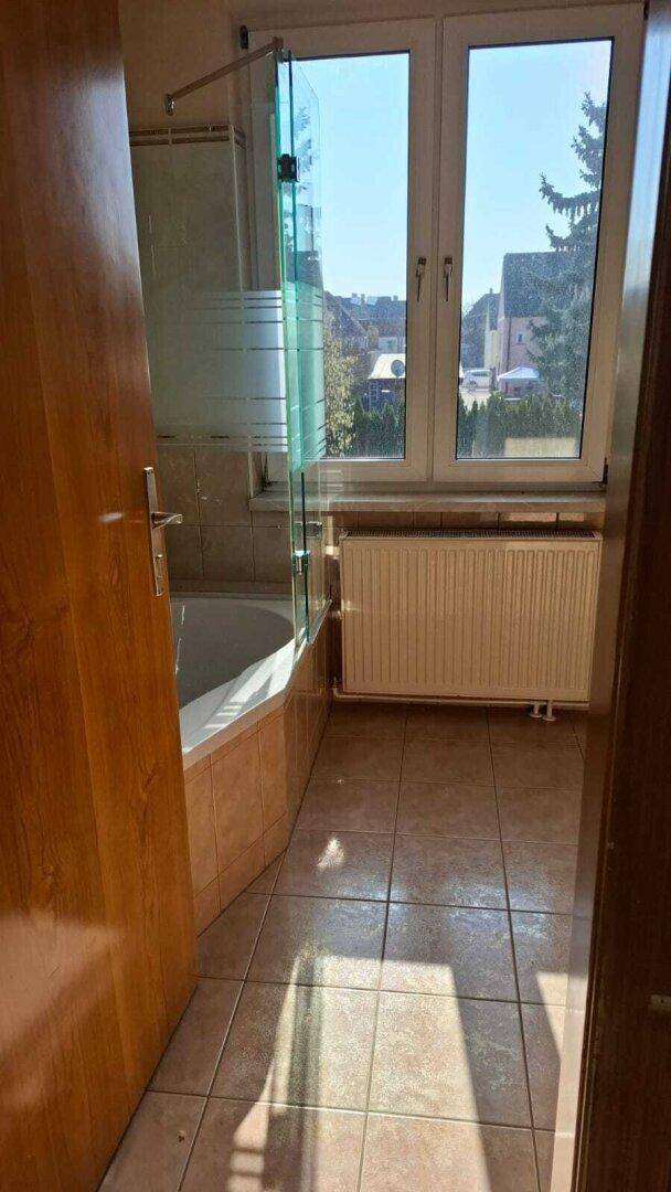 Badezimmer mit Badewanne, Duschwand aus Glas, Fenster und Heizkörper.