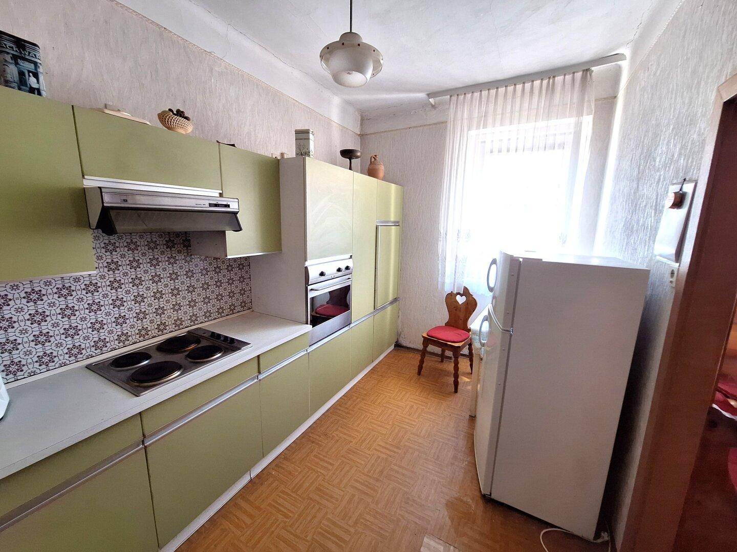 Helle Küche mit grünen Schränken, einem Kühlschrank und einem Fenster mit Vorhängen.